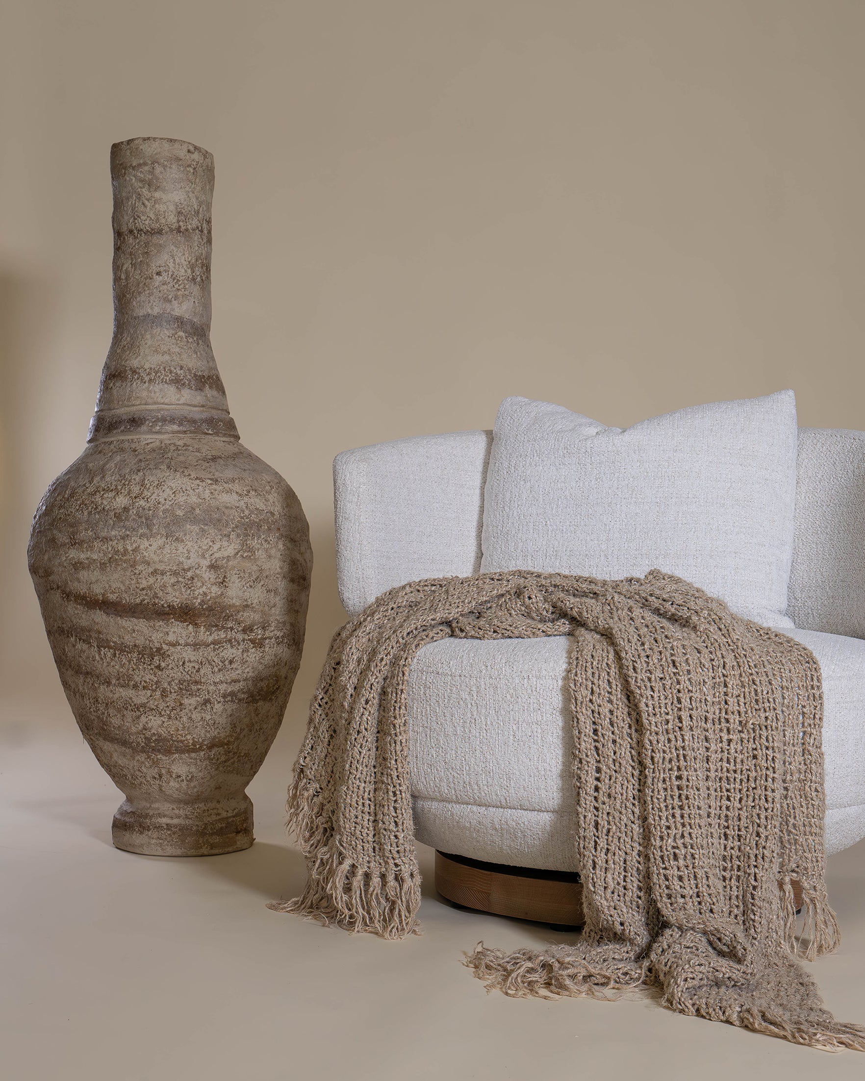 A cosy handwoven taupe throw blanket and matching cushion styled on a curved cream occasional chair, creating a warm and inviting space.