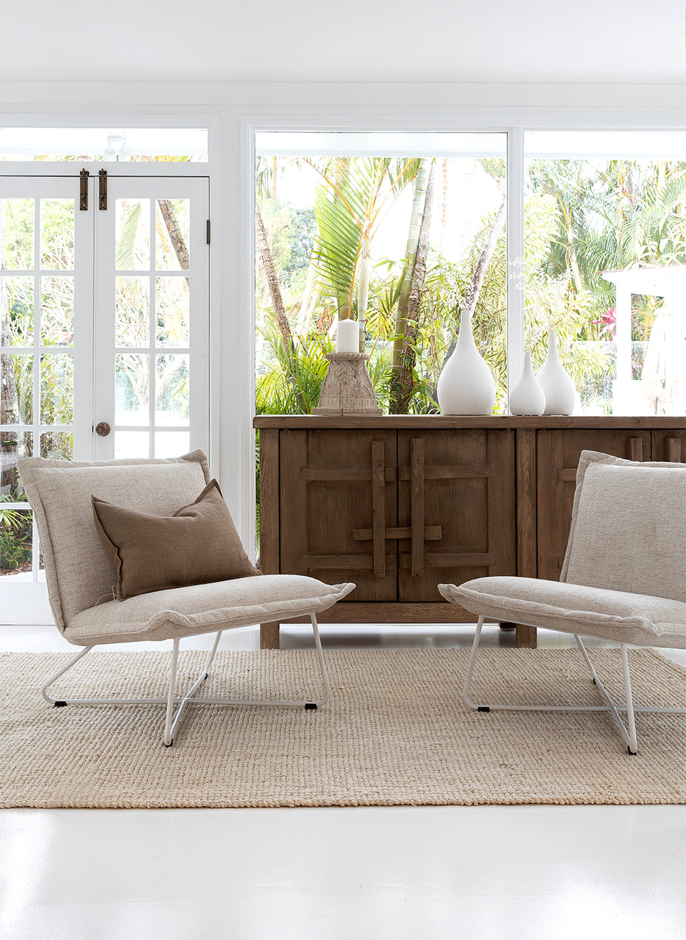 Bright and airy living space by KULALA Living, featuring neutral-toned lounge chairs, a rustic wooden sideboard, and textured décor with lush garden views