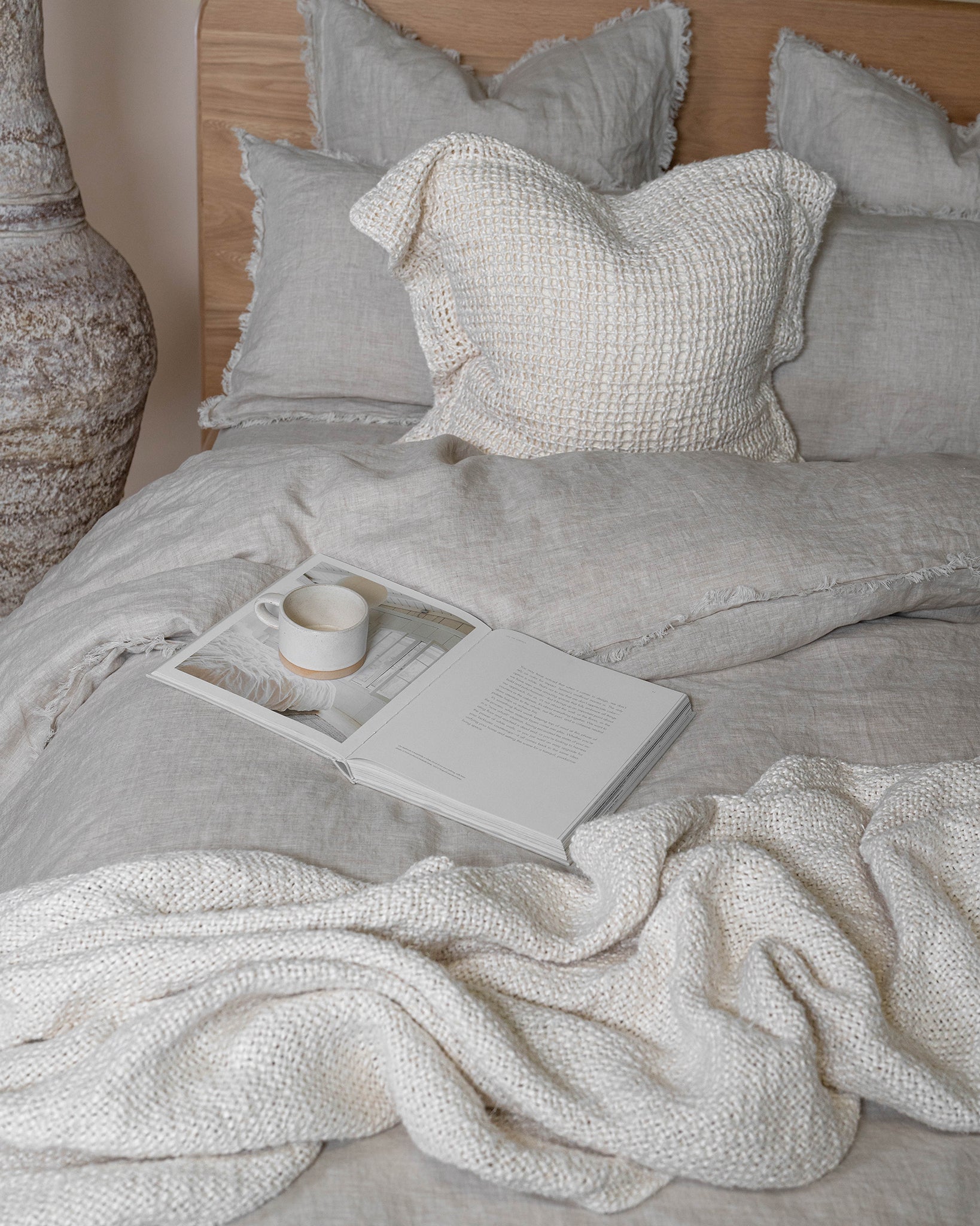 A serene bedroom setting featuring soft linen bedding in neutral tones, complemented by an ivory handwoven cushion with an open-weave texture and a cosy knitted throw. A ceramic cup and an open book add to the relaxed ambiance