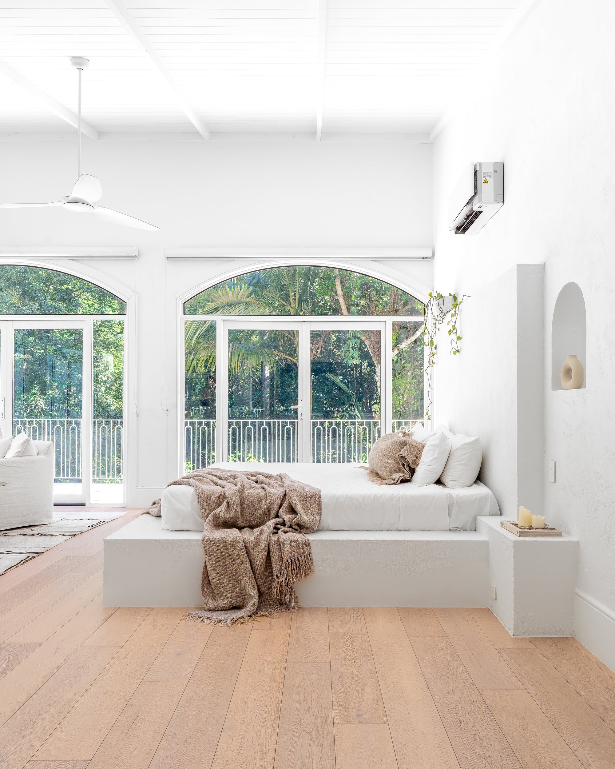 Serene minimalist bedroom featuring a white plaster bed, soft neutral-toned textiles, and large arched windows with lush green views, creating a tranquil retreat