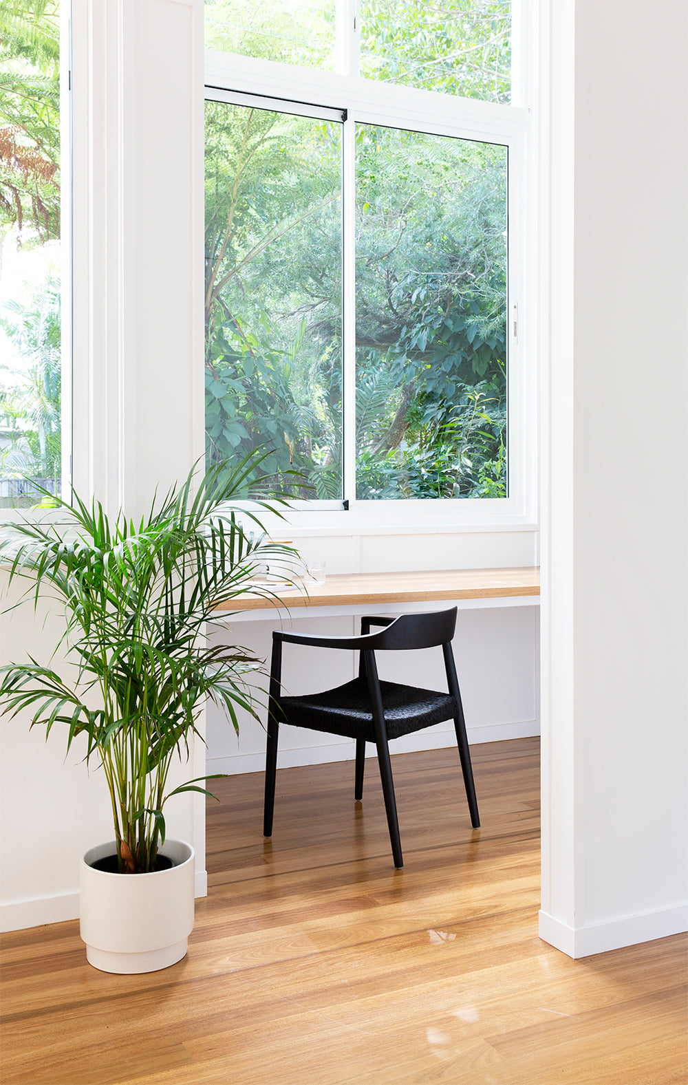 Bright home office with a sleek black woven chair, natural wood desk, and large window overlooking lush greenery. A potted plant adds a fresh, organic touch to the minimalist space