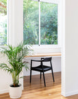 Bright home office with a sleek black woven chair, natural wood desk, and large window overlooking lush greenery. A potted plant adds a fresh, organic touch to the minimalist space