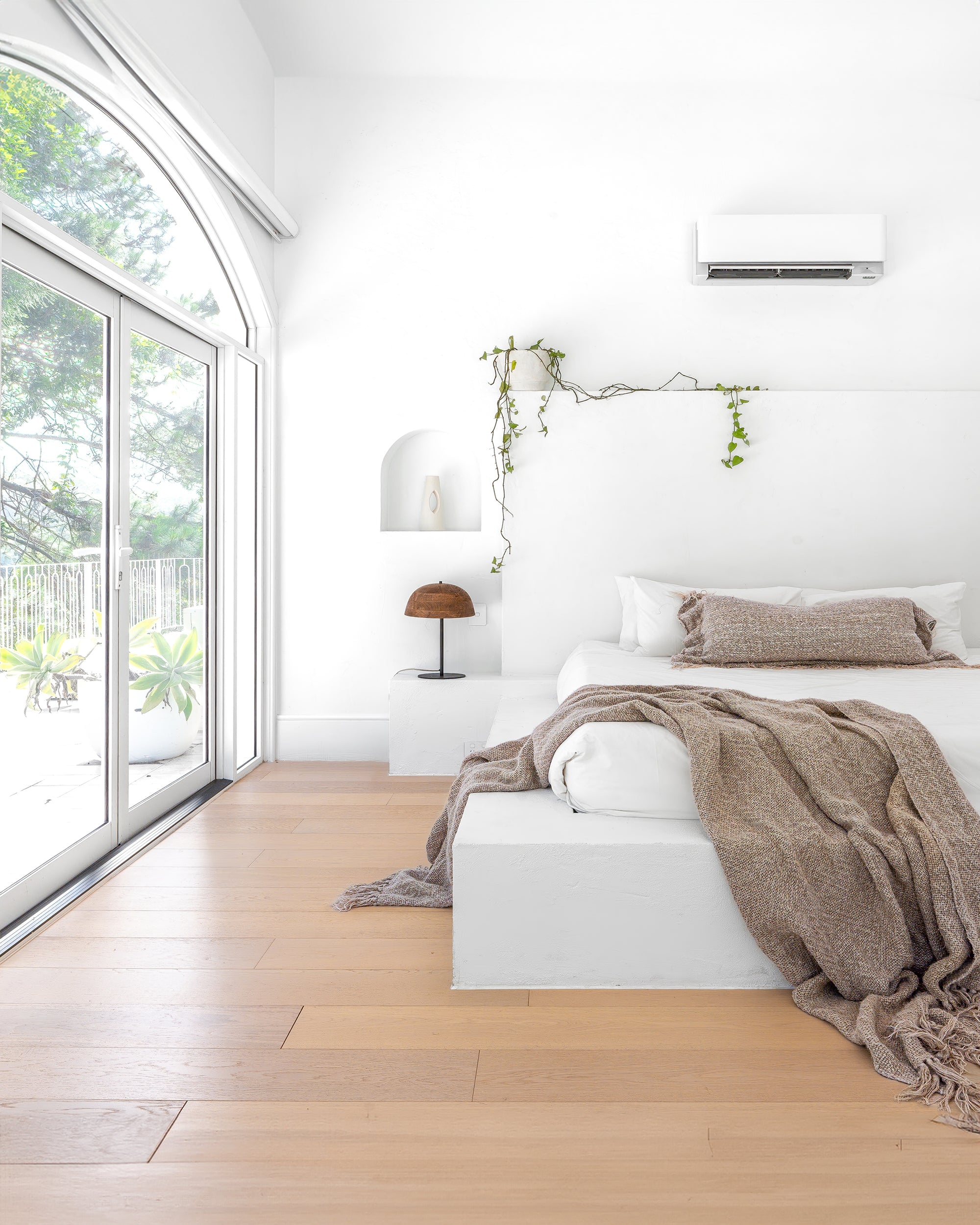 Minimalist bedroom with a white plaster bed, earthy-toned textured bedding, and soft natural light streaming through arched windows, creating a serene retreat