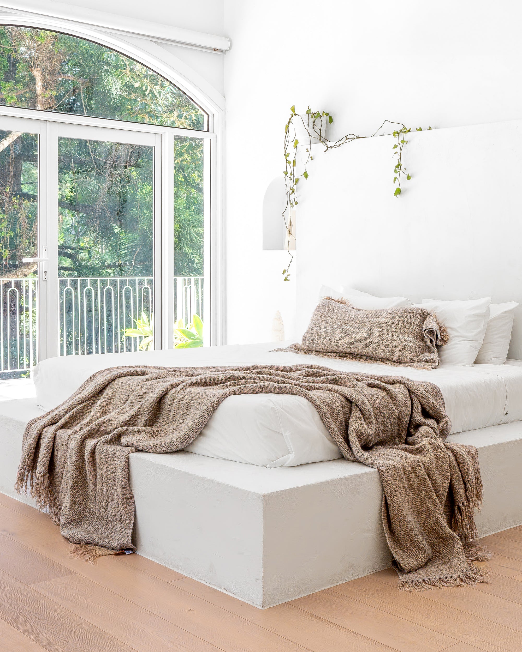 Close-up of a serene minimalist bedroom featuring a white plaster bed with soft, textured bedding in earthy tones, complemented by a cascading vine and natural light from arched windows