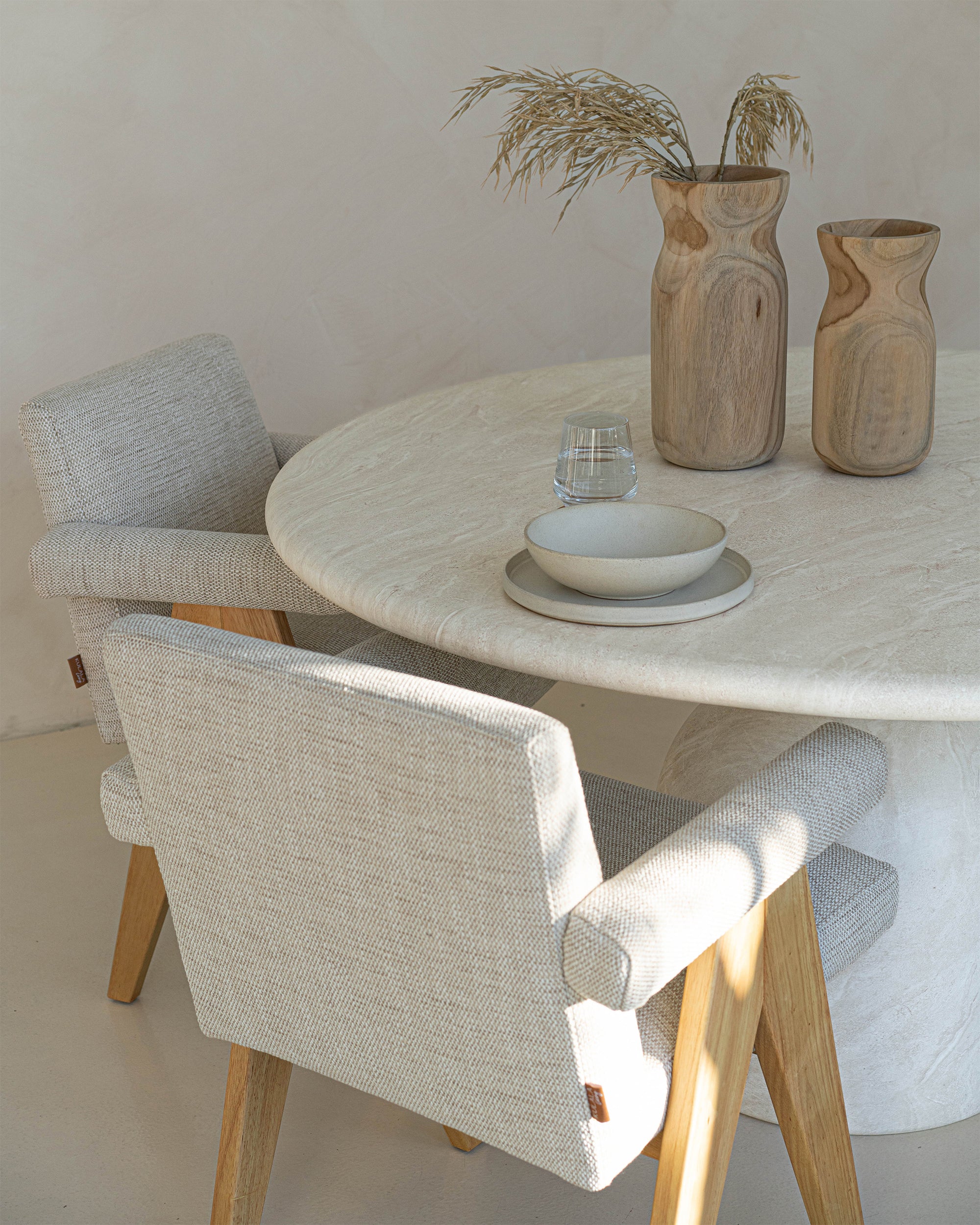 Elegant dining setup featuring modern oak dining chairs with neutral linen upholstery and cushioned armrests, paired with a round travertine table and wooden decorative vases