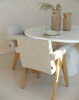 Elegant dining setup featuring modern dining chairs with neutral linen upholstery and cushioned armrests, paired with a round travertine table and wooden decorative vases