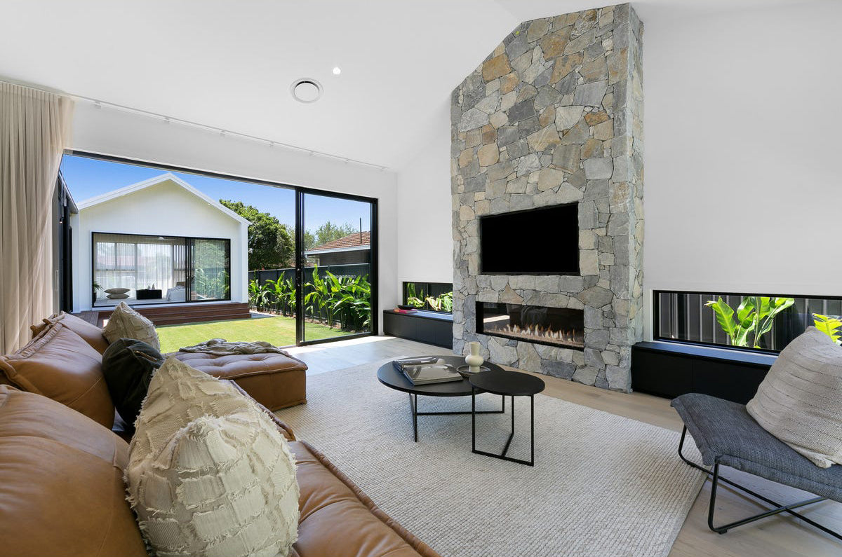 Stylish open-plan living space by KULALA Living, featuring a stone fireplace, leather lounge, textured cushions, and seamless indoor-outdoor flow with lush greenery views