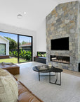 Stylish open-plan living space by KULALA Living, featuring a stone fireplace, leather lounge, textured cushions, and seamless indoor-outdoor flow with lush greenery views