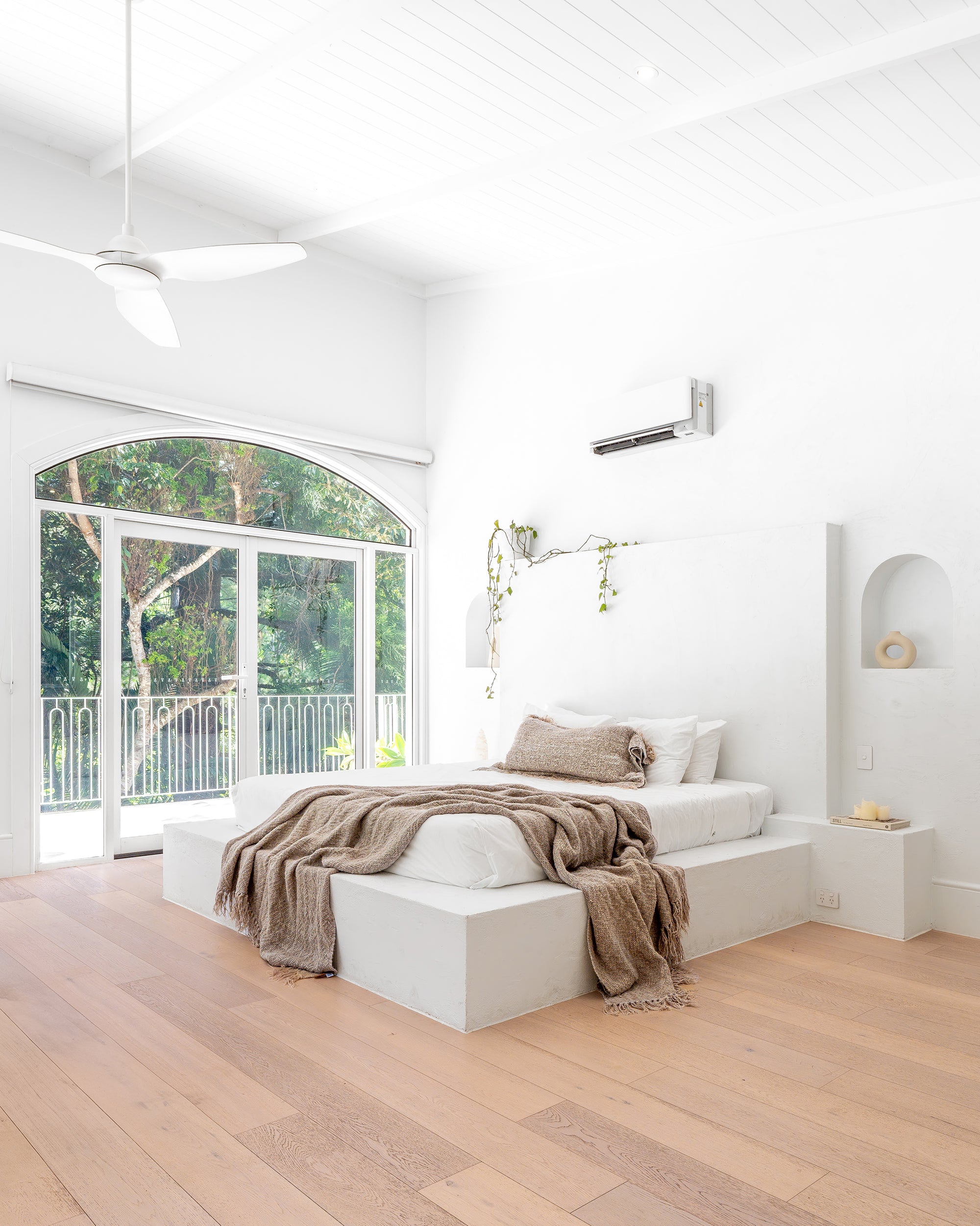 Bright and airy minimalist bedroom with a white plaster bed, soft natural-toned bedding, and large arched windows offering lush green outdoor views, creating a tranquil retreat