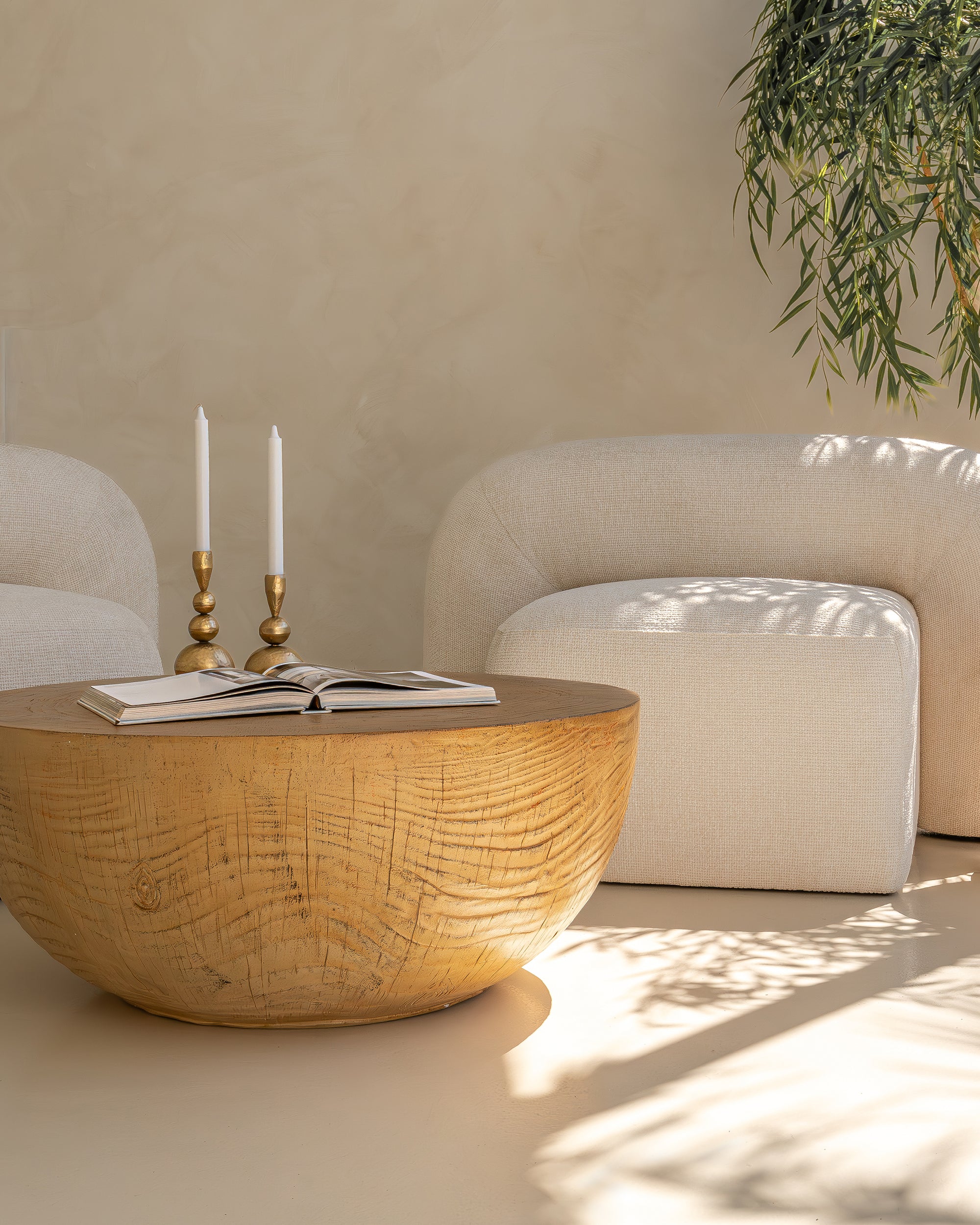 Round coffee table with a textured woodgrain finish, styled with an open book and sculptural brass candlesticks, set between two cream boucle armchairs in a serene, sunlit space