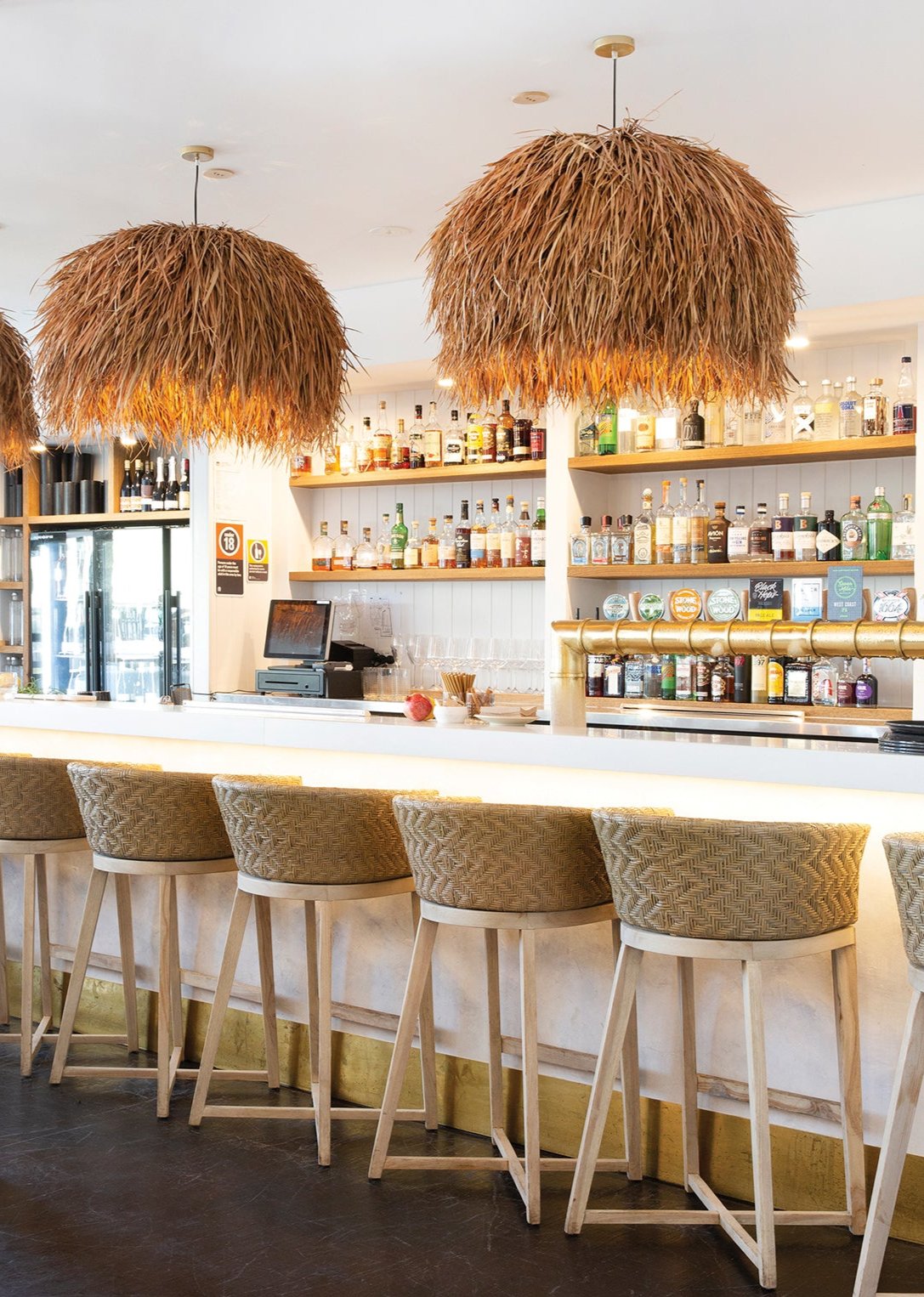 Stylish hospitality interior featuring a row of natural rattan bar stools with woven seats and light wood frames, paired with statement grass pendant lights over a sleek white bar counter