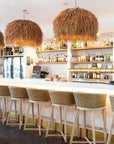 Stylish hospitality interior featuring a row of natural rattan bar stools with woven seats and light wood frames, paired with statement grass pendant lights over a sleek white bar counter