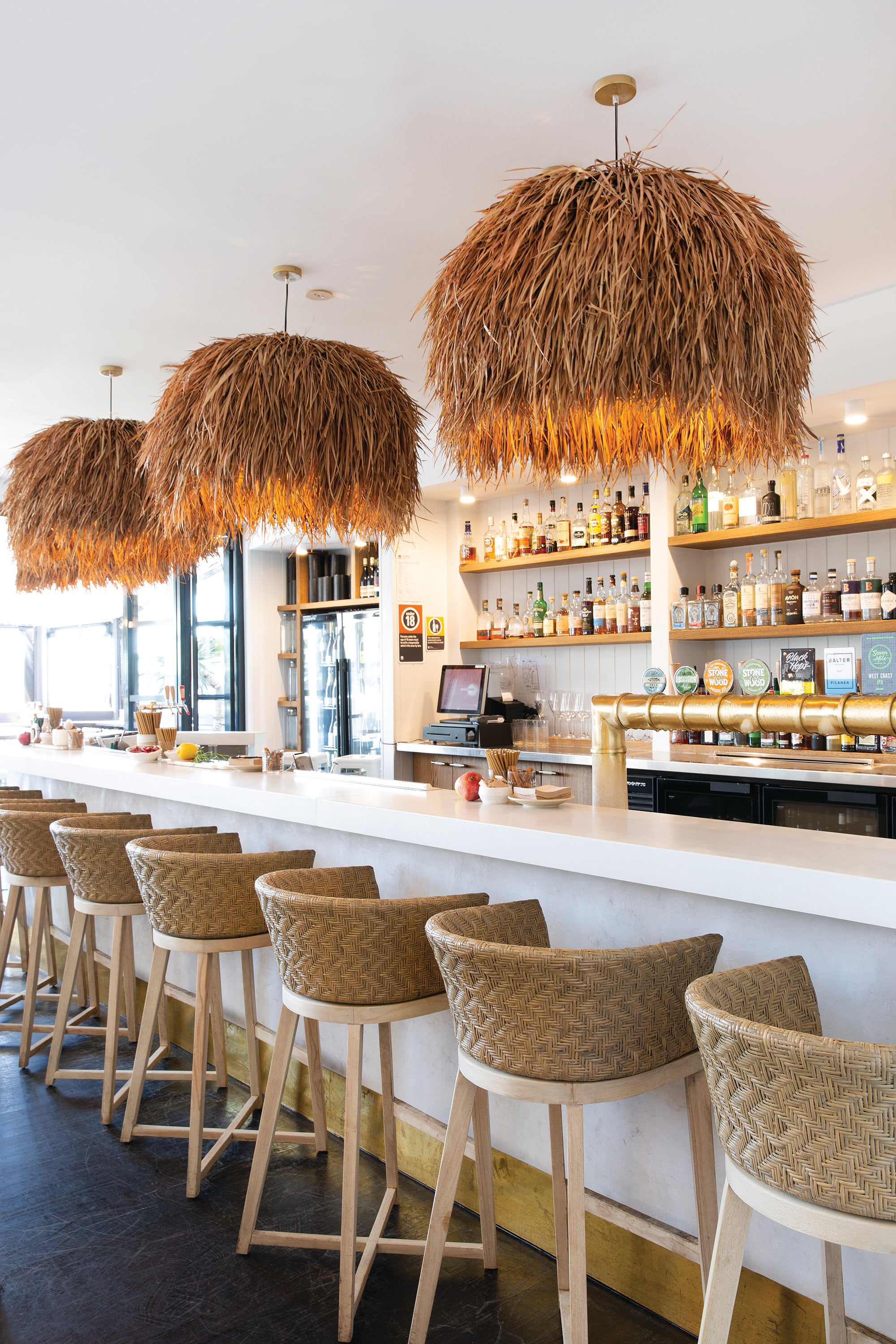 Stylish hospitality interior featuring a row of natural rattan bar stools with woven seats and light wood frames, paired with statement grass pendant lights over a sleek white bar counter