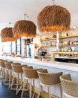 Stylish hospitality interior featuring a row of natural rattan bar stools with woven seats and light wood frames, paired with statement grass pendant lights over a sleek white bar counter