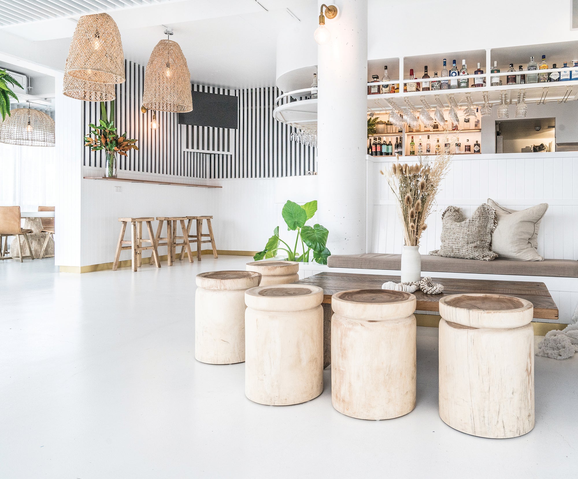 Coastal-inspired bar setting featuring handcrafted natural timber stools, woven pendant lights, and a relaxed seating area with earthy textures and neutral tones