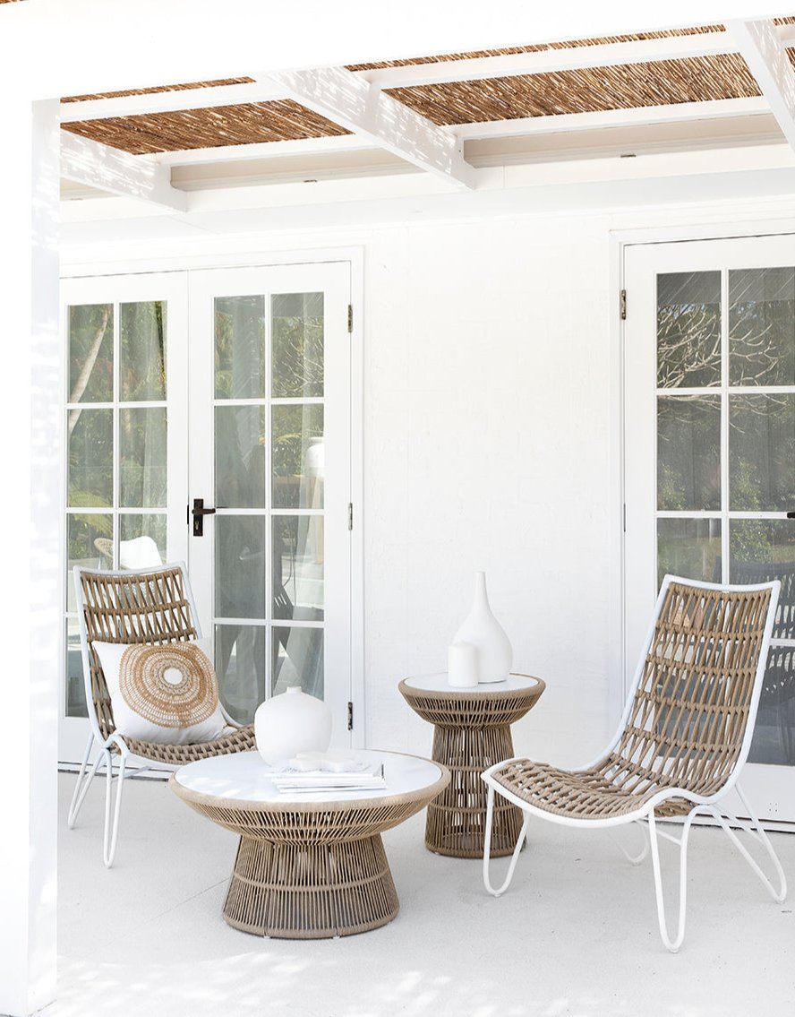 Outdoor lounge setting featuring woven rope chairs with white metal frames and matching side tables, styled with neutral cushions and ceramic decor under a pergola