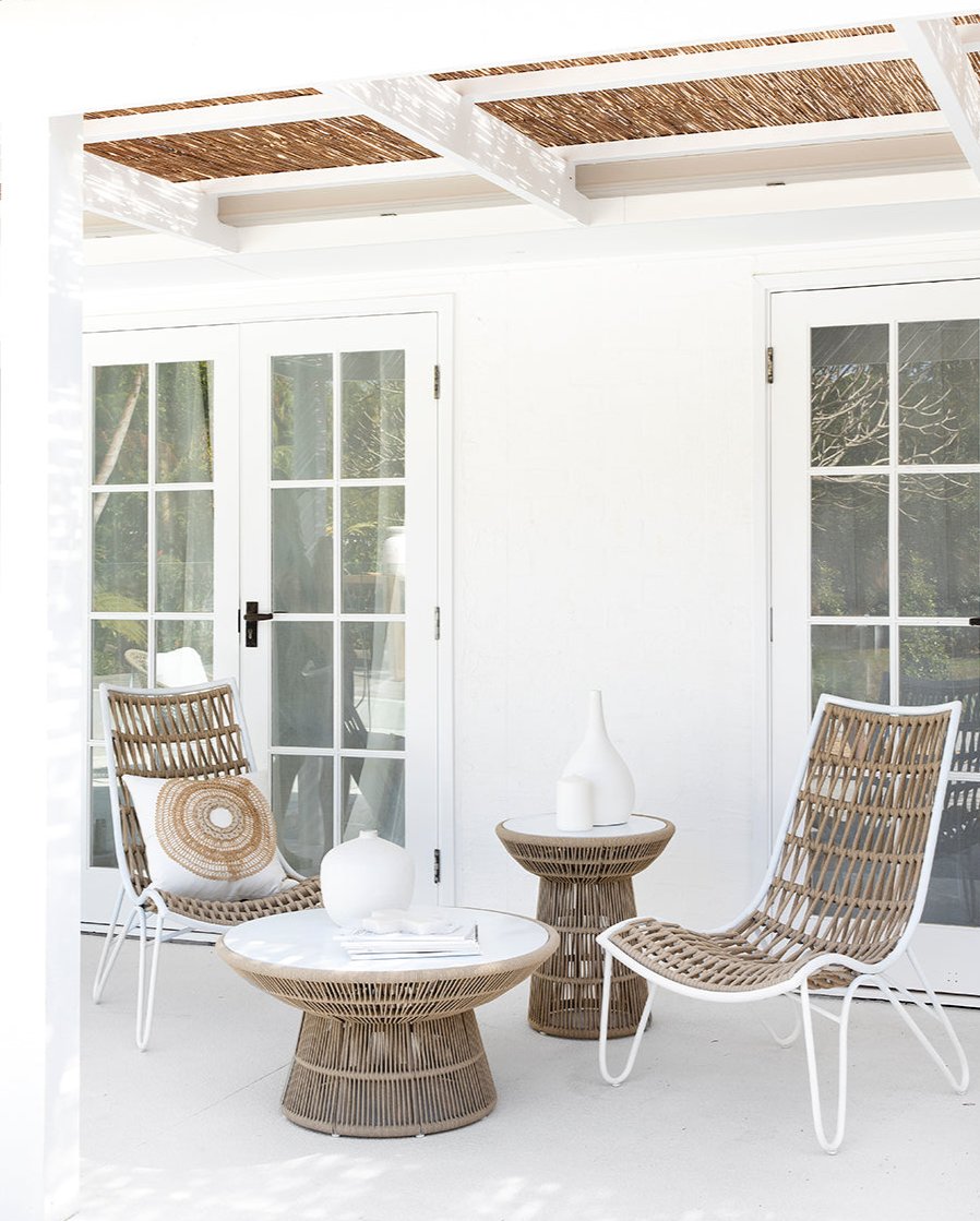 Outdoor lounge setting featuring woven rope chairs with white metal frames and matching side tables, styled with neutral cushions and ceramic decor under a pergola