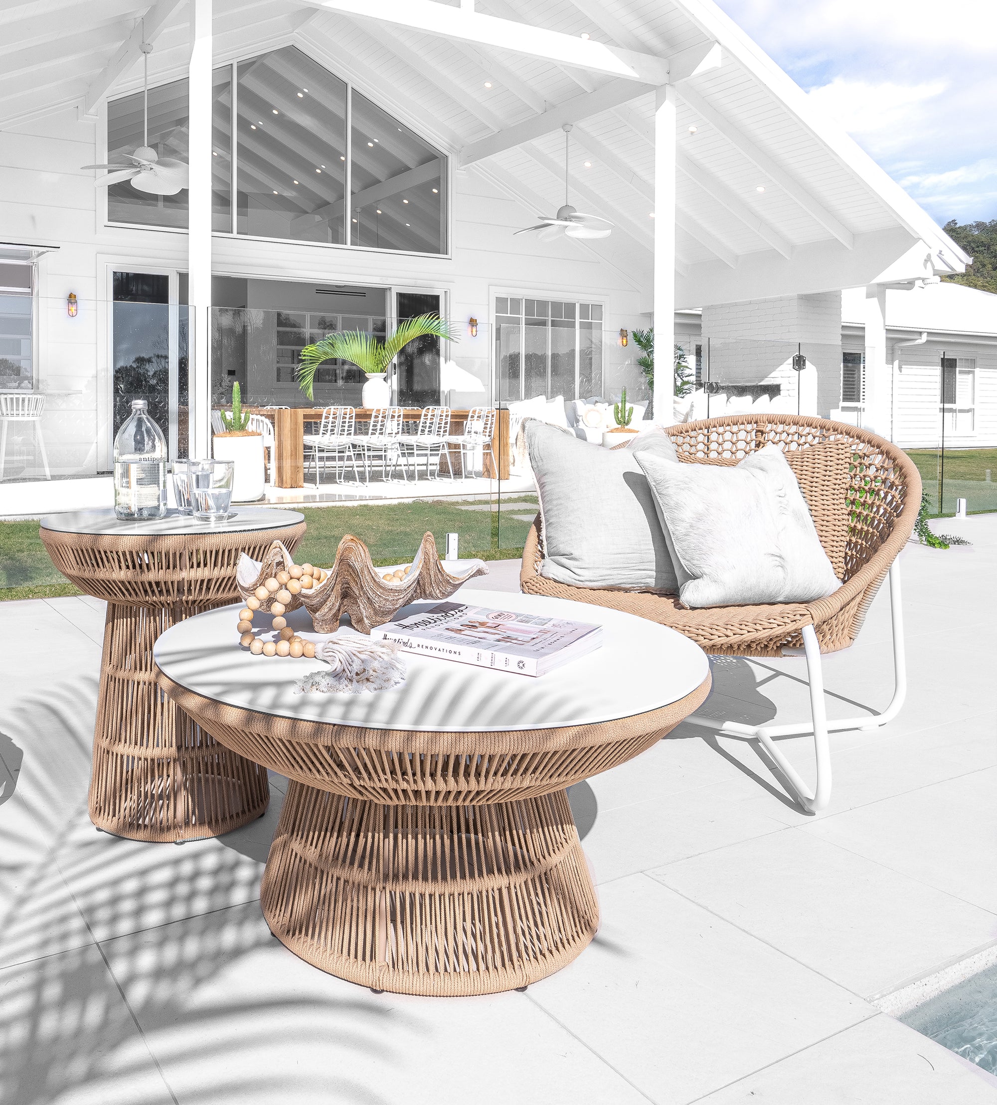 Luxurious outdoor lounge setting featuring woven rope chairs with white metal frames and matching round tables, styled with neutral cushions and coastal decor in a poolside alfresco space