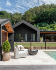 Outdoor setting featuring a contemporary swivel chair and a round black side table by the pool, styled with a ceramic mug and book, set against a black timber-clad home and lush greenery.