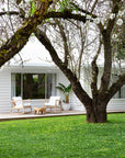 Outdoor deck with two natural wood rocking chairs featuring white cushions, accompanied by a rustic timber stool and a potted tropical plant. Set against a white weatherboard house with lush green lawn and blossoming tree branches in the foreground.