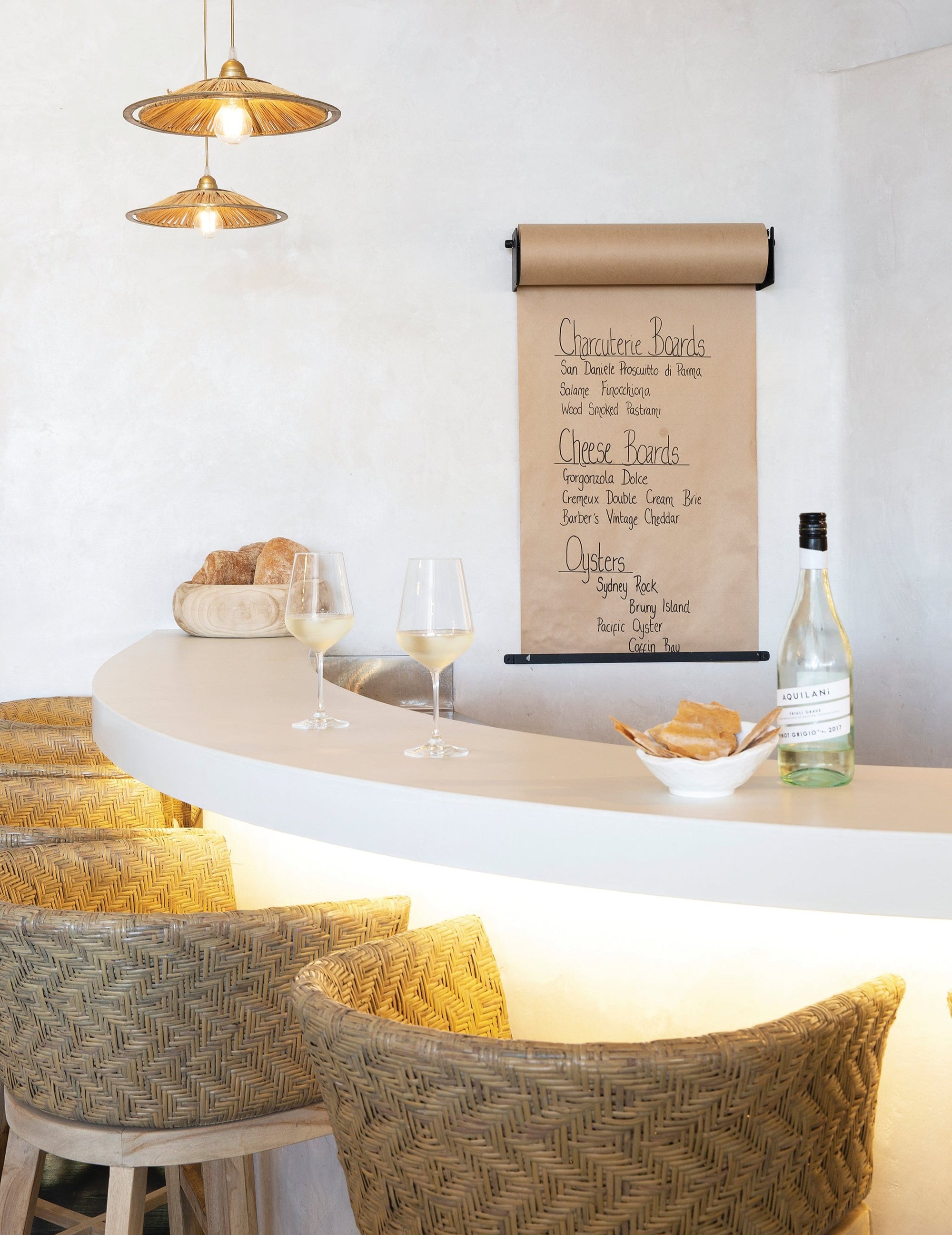 Coastal-inspired bar setting featuring woven rattan bar stools, a curved white counter, pendant lighting, and a handwritten menu on kraft paper, creating a relaxed dining ambiance