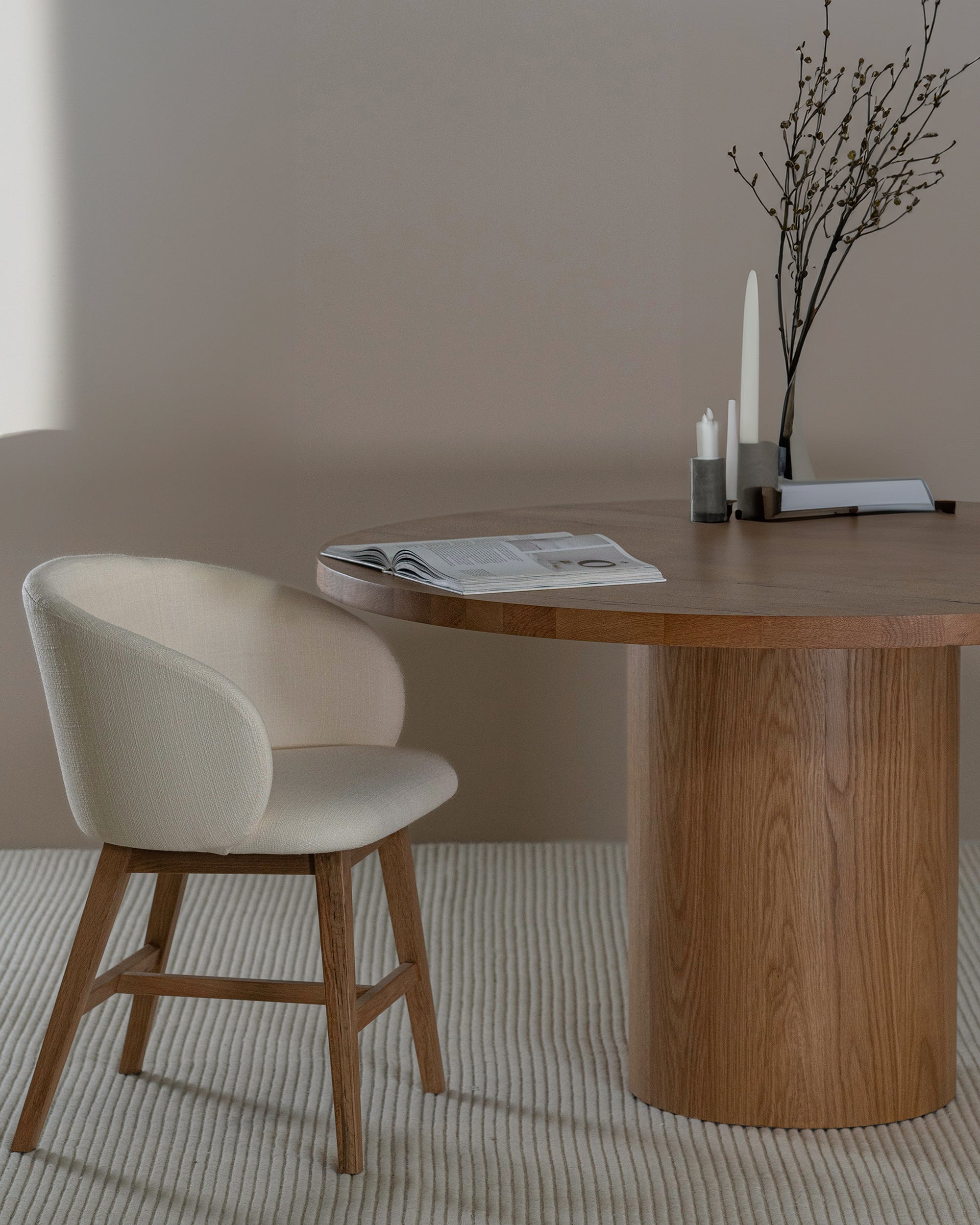 Elegant round oak pedestal dining table paired with a curved, upholstered dining chair in a soft neutral fabric, styled with minimalist decor