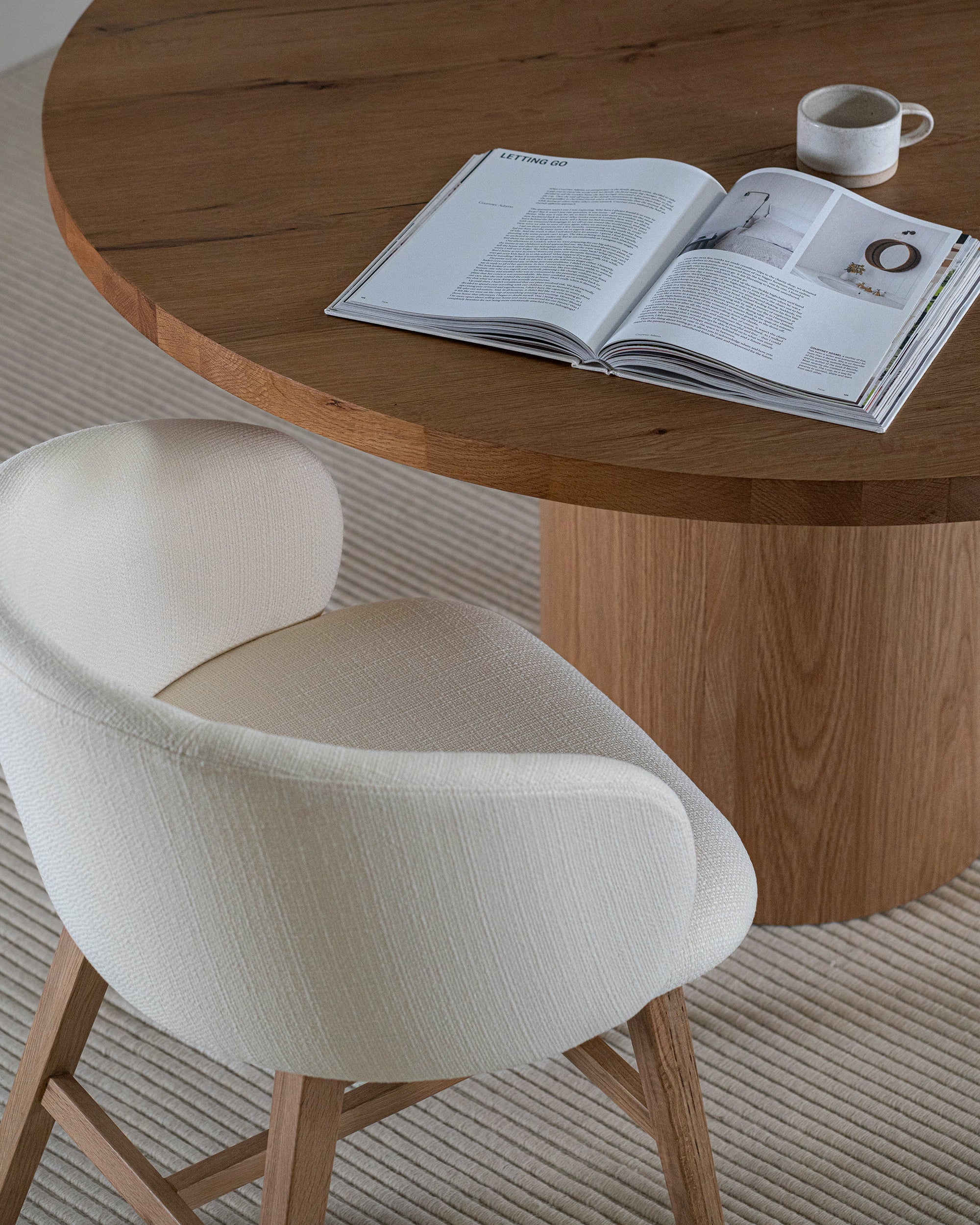 Elegant round oak pedestal dining table paired with a curved, upholstered dining chair in a soft neutral fabric, styled with minimalist decor