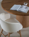 Elegant round oak pedestal dining table paired with a curved, upholstered dining chair in a soft neutral fabric, styled with minimalist decor