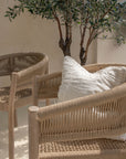 Close-up of KULALA Living teak dining chairs with woven rope detailing, styled with a textured linen cushion in a sunlit, neutral-toned space.