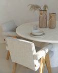 Elegant dining setting featuring textured upholstered chairs with oak legs, a round travertine dining table, and hand-carved teak vases styled with dried foliage