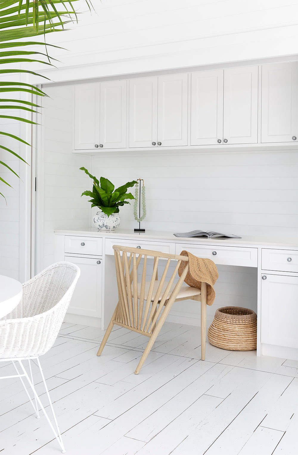 Kulala Living bright coastal home office featuring white cabinetry, a built-in desk, and a lightwood spindle chair with a woven sunhat draped over the back. A potted green plant and decorative accents add warmth to the crisp, airy space