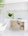 Kulala Living bright coastal home office featuring white cabinetry, a built-in desk, and a lightwood spindle chair with a woven sunhat draped over the back. A potted green plant and decorative accents add warmth to the crisp, airy space