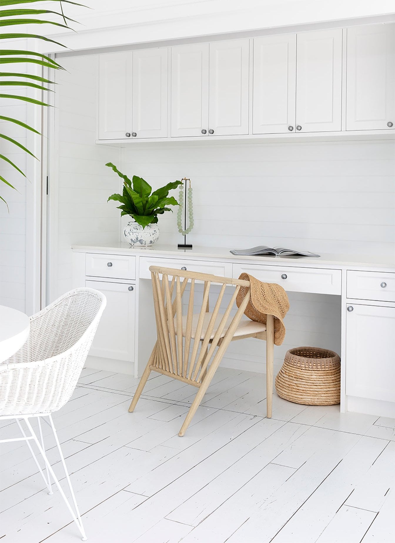 KULALA Living Bright coastal home office featuring white cabinetry, a built-in desk, and a lightwood spindle chair with a woven sunhat draped over the back. A potted green plant and decorative accents add warmth to the crisp, airy space