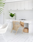 KULALA Living Bright coastal home office featuring white cabinetry, a built-in desk, and a lightwood spindle chair with a woven sunhat draped over the back. A potted green plant and decorative accents add warmth to the crisp, airy space