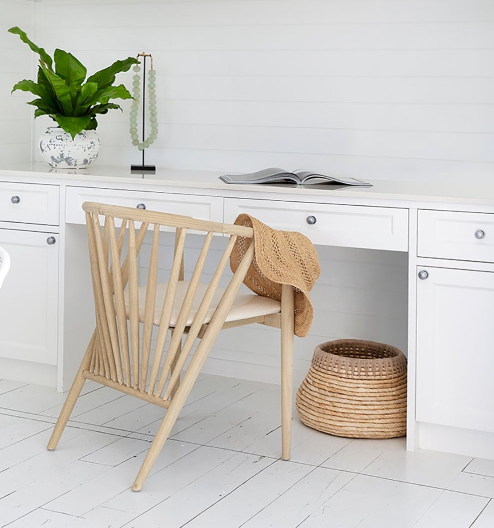 KULALA Living Bright coastal home office featuring white cabinetry, a built-in desk, and a lightwood spindle chair with a woven sunhat draped over the back. A potted green plant and decorative accents add warmth to the crisp, airy space