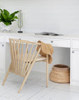 KULALA Living Bright coastal home office featuring white cabinetry, a built-in desk, and a lightwood spindle chair with a woven sunhat draped over the back. A potted green plant and decorative accents add warmth to the crisp, airy space