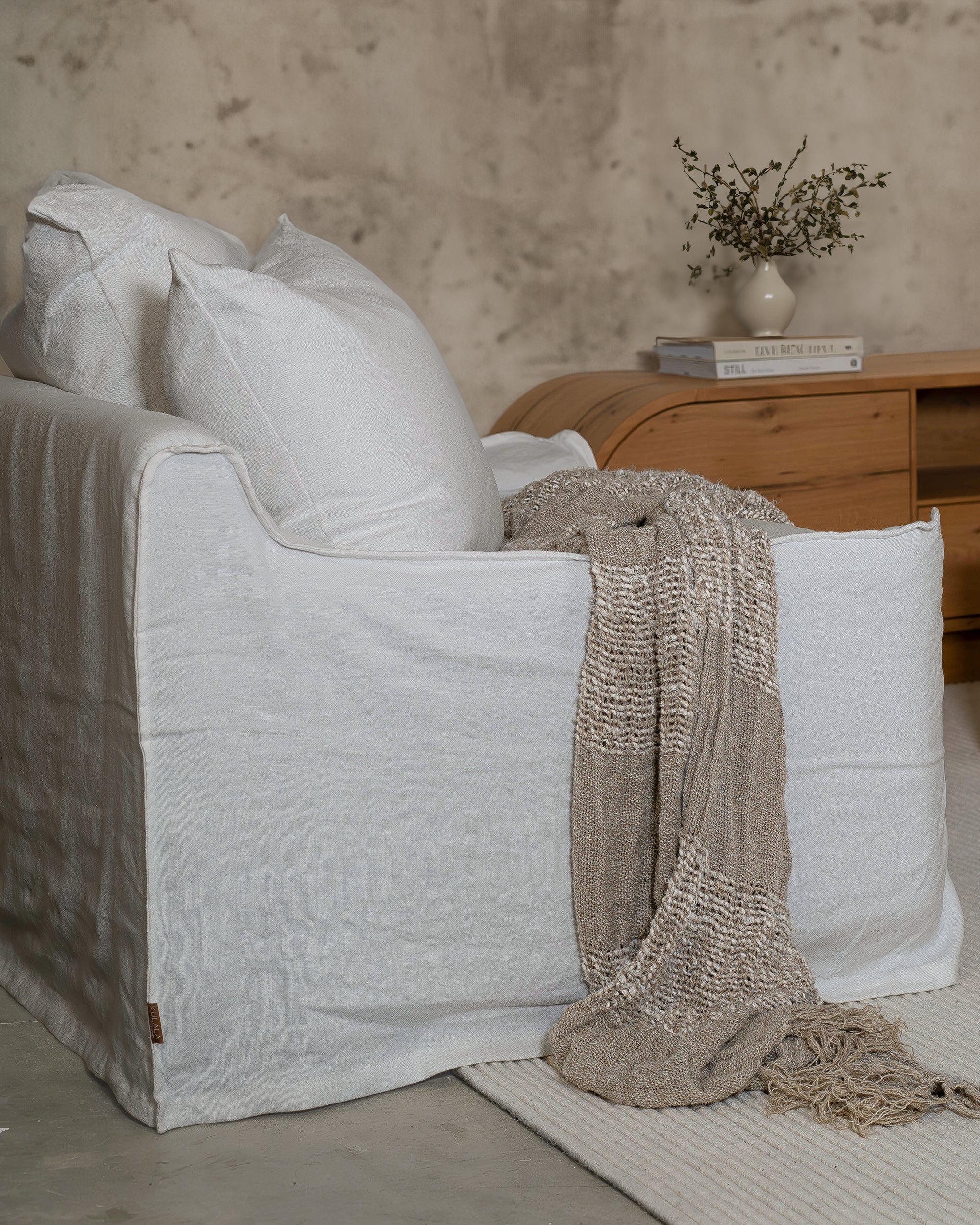 KULALA Living white linen slipcover sofa styled with textured cushions and a neutral throw, creating a relaxed and inviting living space with a natural oak TV unit in the background