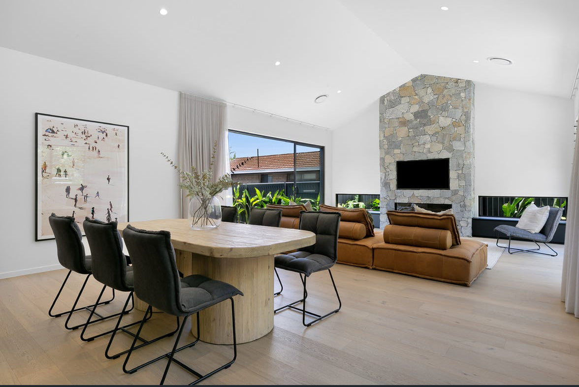 Contemporary dining room featuring black cushioned dining chairs with a sleek metal frame, paired with a rustic round wooden table. The open-plan space includes a stone fireplace, tan leather sofas, and natural light from large windows