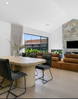 Contemporary dining room featuring black cushioned dining chairs with a sleek metal frame, paired with a rustic round wooden table. The open-plan space includes a stone fireplace, tan leather sofas, and natural light from large windows