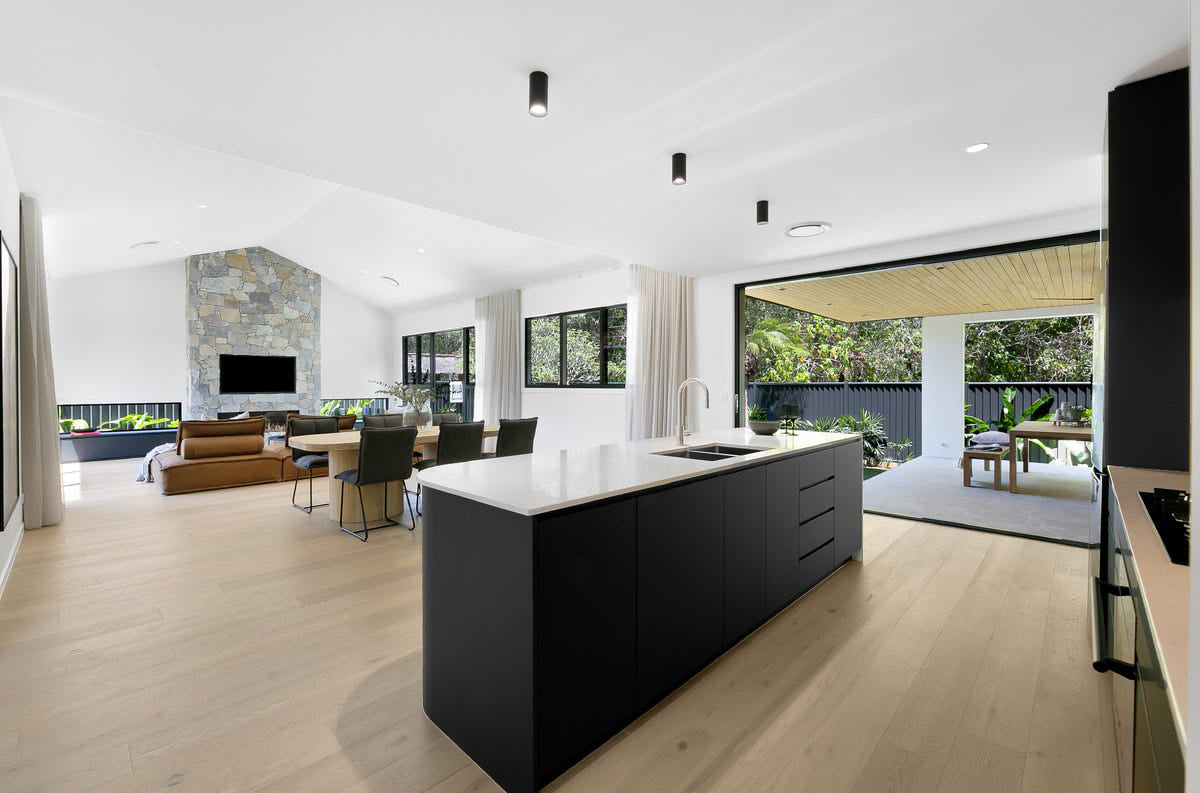 Modern open-plan kitchen with a sleek black island and light timber flooring, seamlessly flowing into the dining and living area. Featuring a stone fireplace, black cushioned dining chairs, and large sliding doors opening to an outdoor entertaining space with lush greenery