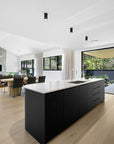 Modern open-plan kitchen with a sleek black island and light timber flooring, seamlessly flowing into the dining and living area. Featuring a stone fireplace, black cushioned dining chairs, and large sliding doors opening to an outdoor entertaining space with lush greenery