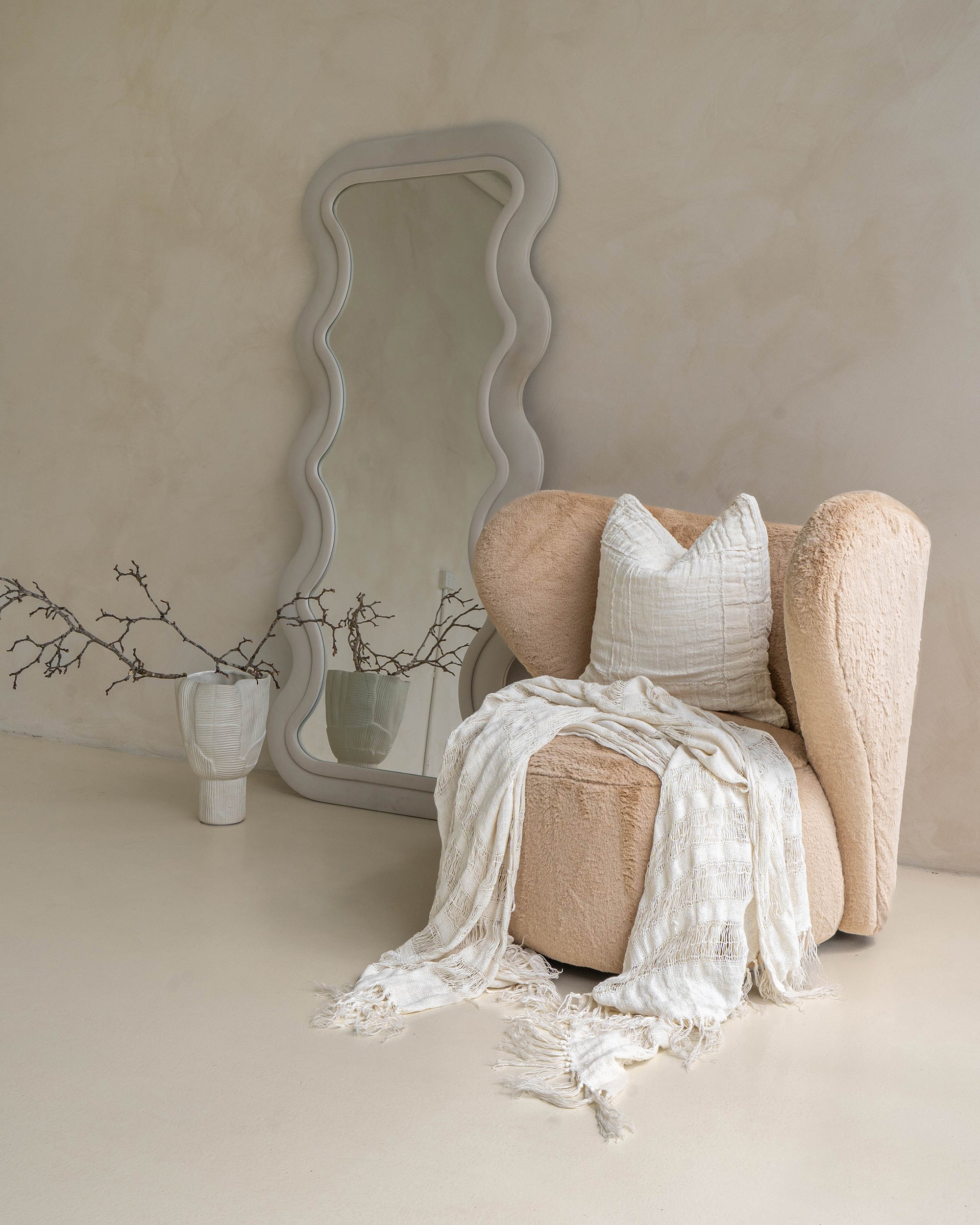 Elegant neutral-toned lounge setting featuring a textured armchair, handwoven white linen throw with delicate fringe, and a matching cushion, styled beside a sculptural wavy mirror and ceramic vases