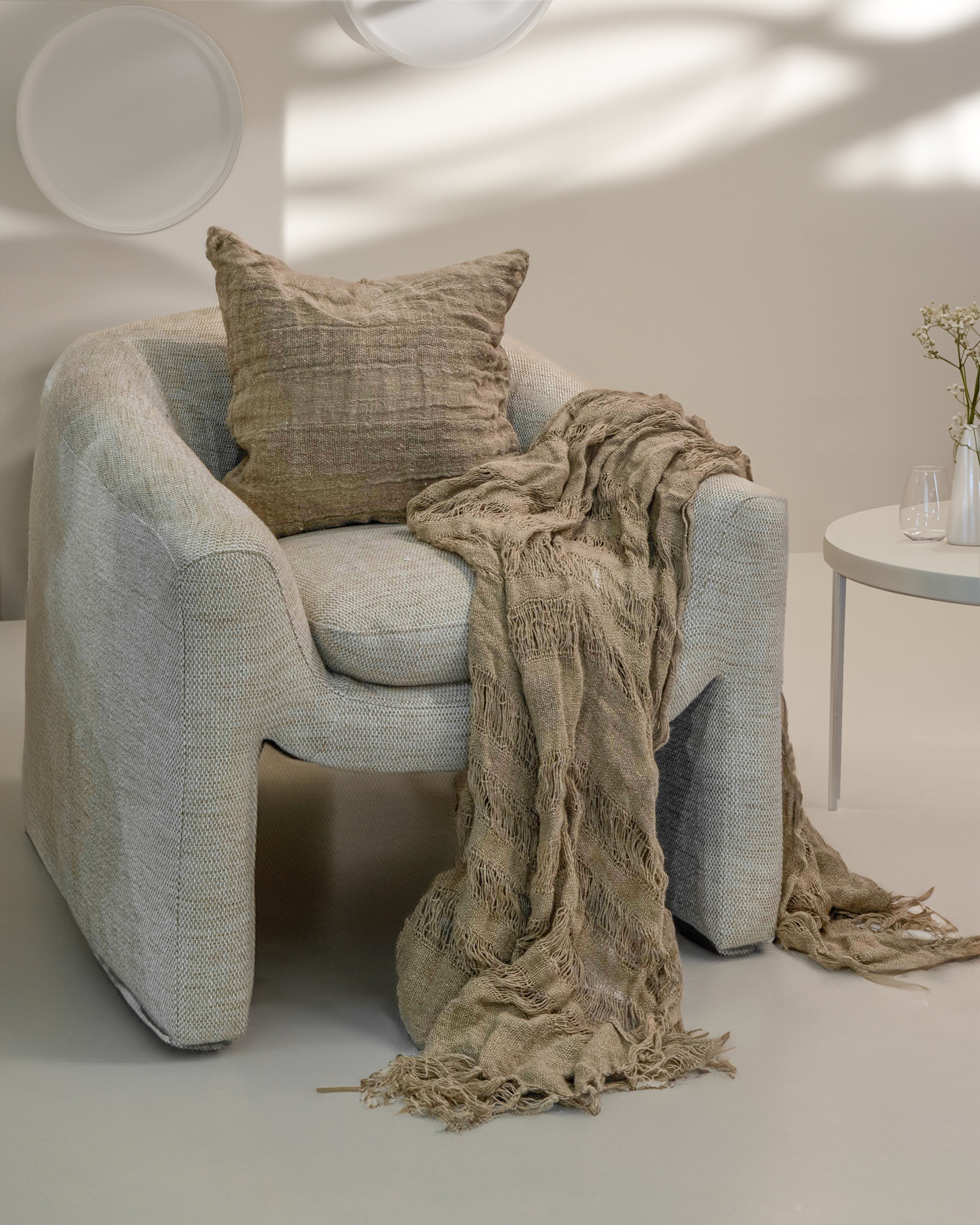 Earth-toned textured linen throw with delicate fringe detail draped over a modern neutral armchair, paired with a matching cushion, creating a relaxed and inviting aesthetic in a softly lit space