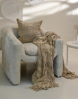 Earth-toned textured linen throw with delicate fringe detail draped over a modern neutral armchair, paired with a matching cushion, creating a relaxed and inviting aesthetic in a softly lit space